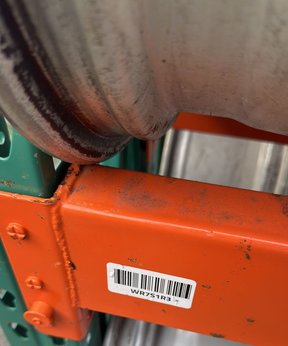 Outdoor orange pallet racking holding bare wheels, with a barcode label on the upright reading WR7S1R3