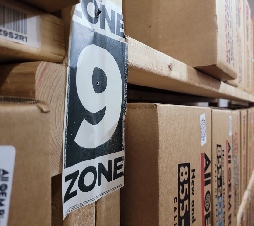 A warehouse rack holding boxed wheels. A hanging paper sign reads ZONE 9 for a person; on the shelf beside it a printed barcode label reads Z9S2R1 for a scanner